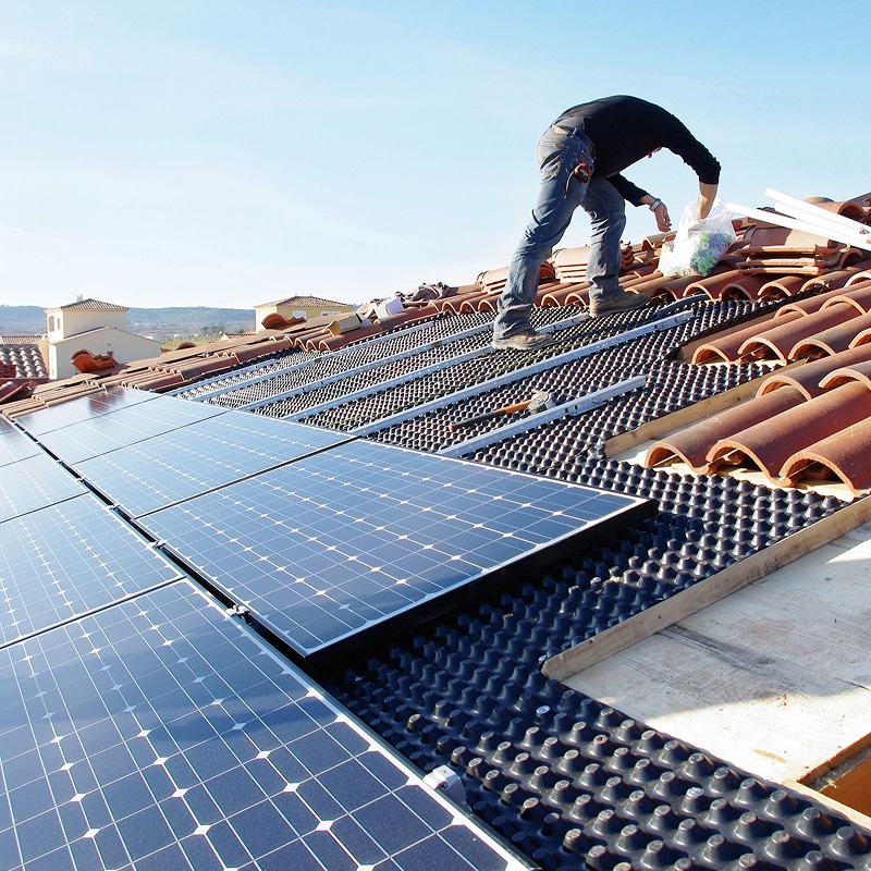 Installation Solaire : Pose de Panneaux Photovoltaïques sur Toit Technicien installant des panneaux photovoltaïques sur un toit de tuiles. La membrane noire et les rails de fixation soutiennent la structure solaire.